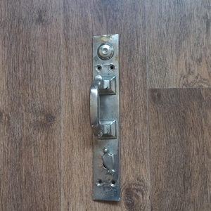 Op de afbeelding: Een vintage zilverkleurige deurklink en plaat, verticaal gemonteerd. De hendel is een gebogen staaf en de plaat heeft decoratieve vierkante elementen en een sleutelgat. De hardware staat tegen een achtergrond van houtnerf.