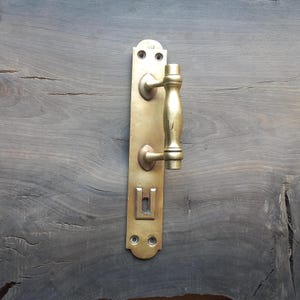 May include: A vintage brass door handle set. The handle has a vertical rectangular backplate with a keyhole and two screw holes. The handle itself is a curved, cylindrical shape. The brass has a warm, aged patina.