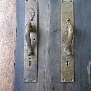 May include: Two antique brass door handles with keyholes and ornate detailing. The handles are mounted on a weathered, dark gray wooden surface, showcasing a vintage aesthetic. One handle features a leaf design.
