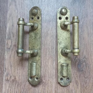 May include: Two antique brass door handles with a weathered finish. Each handle features a long, rectangular backplate with decorative detailing, a cylindrical handle, and a smaller, lower cylindrical element. The handles are arranged side by side on a wooden surface.