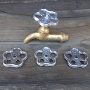 Puede incluir: Un grifo antiguo de latón con un mango plateado y tres mangos plateados adicionales. El grifo está colocado sobre una superficie de madera envejecida, mostrando una estética vintage. Los mangos tienen un diseño en forma de flor.