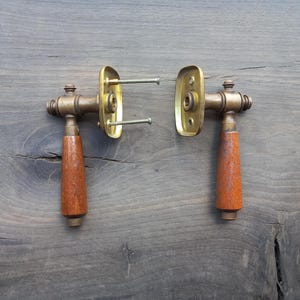 May include: Two antique door handles with brass hardware and wooden grips. The handles are rectangular with rounded edges and are attached to brass plates with screws. The wooden grips are a reddish-brown color.
