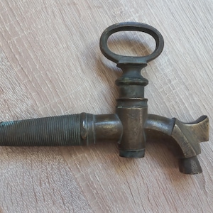 Può includere: Un rubinetto per botte in metallo color bronzo scuro con un beccuccio conico filettato su un lato, un corpo centrale verticale e una maniglia superiore a forma di anello ovale. Il beccuccio si curva verso il basso all'estremità opposta.