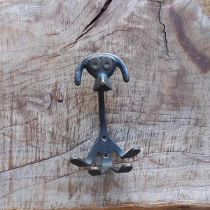 May include: A quirky, dark blue metal wall hook in the shape of a dog. The hook has a dog-like face with two round eyes and a long body with three hooks at the base. It is mounted on a rustic wooden surface.