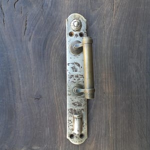 May include: An antique door handle with a distressed, aged appearance. The handle is made of metal with a tarnished, silver finish and a cylindrical brass handle. It is mounted on a dark wooden door.