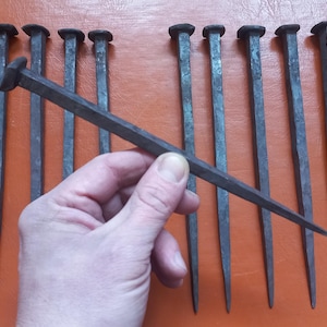 Op de afbeelding: Een verzameling antieke, donkergrijze ijzeren spijkers. Eén spijker wordt in de hand gehouden, waarbij het puntige uiteinde en de vierkante kop te zien zijn. De spijkers zijn geplaatst tegen een oranje achtergrond, wat hun verouderde uiterlijk benadrukt.