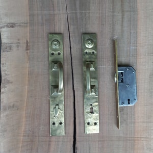 May include: Two brass door handles with keyholes and a matching brass door latch. The handles have a rectangular shape with a decorative design and a lever handle. The latch is a long, thin piece of metal with a keyhole and a small lever.