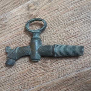 Op de afbeelding: Een antieke bronzen kraan met een ringhandvat en een schroefdraaduitloop. De kraan heeft een hendelachtig ontwerp en is waarschijnlijk voor een vat of container. De kraan staat tegen een houten achtergrond.