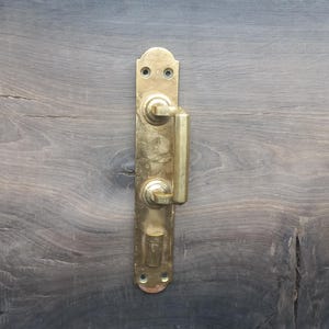 May include: A vintage brass door handle with a vertical rectangular backplate, mounted on a weathered wooden door. The handle features a lever and a small, round knob. The wood grain is visible.