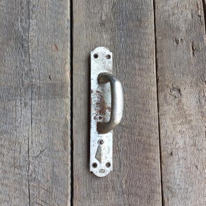 May include: A vintage, weathered door handle with a curved, silver-toned grip. The handle is mounted on a rectangular, aged metal plate with visible rust and screw holes. The background is a rustic wooden door.