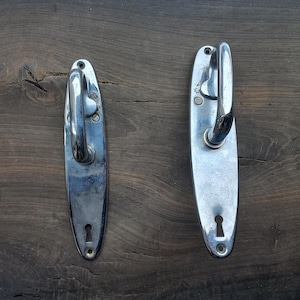 Può includere: Due maniglie per porte argentate con piastre ovali e buchi per le chiavi. Le maniglie hanno un design curvo e sono montate su una superficie in legno.