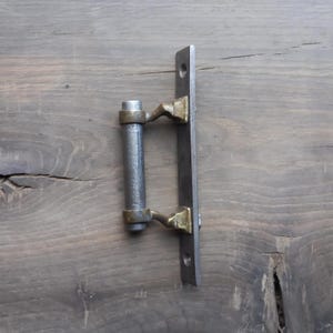 May include: A vintage-style door pull featuring a dark grey metal handle and a rectangular mounting plate. The handle is cylindrical with brass-coloured detailing. The mounting plate has two pre-drilled holes for installation.