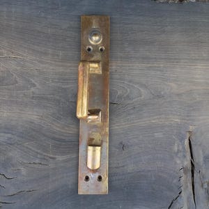 Könnte beinhalten: Ein antiker Türgriff aus Messing mit rechteckiger Grundplatte. Der Griff hat einen Hebel und ein kleines zylindrisches Element. Das Messing weist eine gealterte Optik mit Gebrauchsspuren und Oxidation auf.