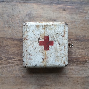 May include: A vintage, rectangular first aid kit with a white, distressed finish and a prominent red cross emblem. The metal case shows signs of age and wear, with rust and discoloration. A metal clasp secures the lid.