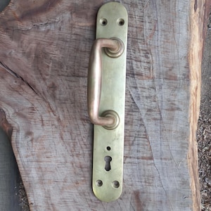 Op de afbeelding: Een vintage messing deurklink met een gebogen handvat en een rechthoekige achterplaat. De klink heeft een warme, gouden tint en is gemonteerd op een verweerd houten oppervlak. Het sleutelgat is zichtbaar eronder.