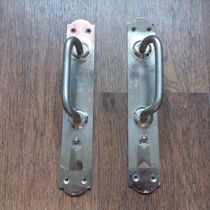 Può includere: Due maniglie per porta in metallo color argento abbinate, ciascuna dotata di una lunga piastra posteriore rettangolare con estremità arrotondate e due fori per le viti in alto e in basso. Una robusta maniglia a U è montata centralmente su ogni piastra.