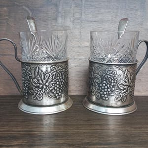 Puede incluir: Dos portavasos plateados vintage con asas, cada uno con una taza de cristal tallado transparente y una cucharita. Los portavasos están decorados con un diseño de uvas y hojas, y están sobre una superficie de madera.