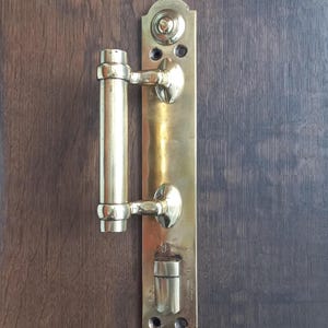 May include: A brass door handle with a vertical rectangular backplate and a cylindrical handle. The handle has a polished finish and is mounted on a dark brown wooden door. The door handle is an antique style.