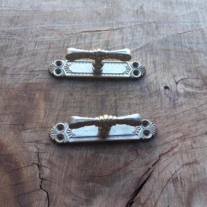 May include: Two vintage brass door handles with an ornate design. Each handle features a textured central grip and is mounted on a rectangular backplate with decorative detailing. The handles are set against a rustic wooden surface.