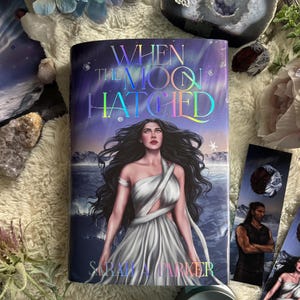 Pode incluir: Um livro intitulado "When the Moon Hatched" com uma mulher em um vestido branco na capa. A capa tem um brilho holográfico com o nome da autora, Sarah A. Parker, na parte inferior. Marcadores com arte semelhante também são visíveis.