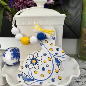 May include: A white ceramic jar with a lid and a blue and yellow bunny rabbit ornament. The bunny is decorated with white flowers and blue and yellow polka dots. The jar has a string of white and yellow beads around it.