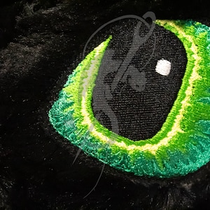 May include: Close-up of an embroidered eye with a black pupil and a bright green iris. The eye is set against a black, textured background, possibly fur or fabric. The eye's details are meticulously stitched.