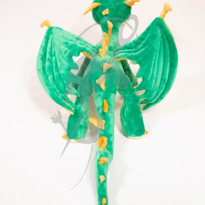 May include: Green plush dragon toy with yellow accents. The dragon has wings, a long tail, and a spiky back. The toy is standing on a white surface. PDF PATTERN.