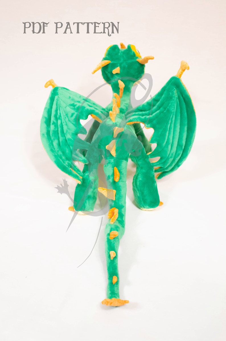 May include: Green plush dragon toy with yellow accents. The dragon has wings, a long tail, and a spiky back. The toy is standing on a white surface. PDF PATTERN.
