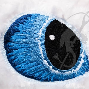 May include: Close-up of an embroidered eye with a black pupil and a bright blue iris. The eye is set against a soft, white, textured background. The embroidery detail is intricate, showcasing the texture of the thread.