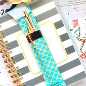 May include: A turquoise and gold pen holder with a mermaid scale pattern. The holder has a black velcro closure and the text "Planner O's" is printed on the bottom. The holder is attached to a gray and white striped planner.