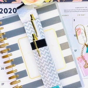 May include: A white and gray chevron patterned pen holder with a black elastic band. The pen holder is attached to a gray and white striped planner with gold rings. The planner has the year 2020 printed on the cover.