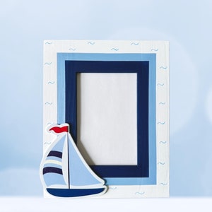 May include: A white wooden picture frame with a blue and white sailboat design. The frame has a blue border and a white background with blue wavy lines.