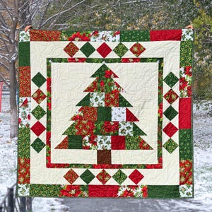 Könnte beinhalten: Eine Weihnachtsdecke mit einem zentralen Baumdesign. Der Baum besteht aus grünem, rotem und weiß gemustertem Stoff. Die Decke hat einen cremefarbenen Hintergrund und ist mit roten, grünen und floral gemusterten Quadraten und Rauten umrandet.