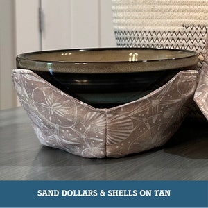 May include: A tan bowl cozy featuring a pattern of sand dollars and shells. The cozy is wrapped around a dark-colored bowl. The text "SAND DOLLARS & SHELLS ON TAN" is at the bottom of the image.