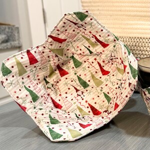 May include: A fabric bowl with a Christmas tree pattern in red, green, and gold on a cream background. The bowl has a square shape with a folded-over edge. The interior is lined with the same festive fabric.