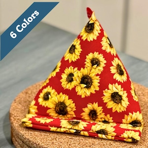 May include: A red fabric item with a sunflower pattern. The item is triangular, with a folded base, and a small red loop at the top. The words "6 Colors" are in a blue banner in the upper left corner. The item is on a cork surface.