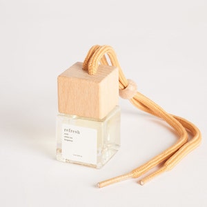 Könnte beinhalten: Eine kleine, klare Glasflasche mit einem Holzdeckel und einer beigefarbenen Schnur. Die Flasche enthält eine Flüssigkeit und trägt ein Etikett mit der Aufschrift "refresh" mit den Duftnoten "Yuzu, weißer Tee, Bergamotte".