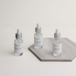 May include: Three glass bottles with silver caps and labels. The labels are white with black text. The bottles are on a gray hexagonal tile. The labels read "Yuzu Leaf", "dusty rose", "kitchen window", "earl grey".