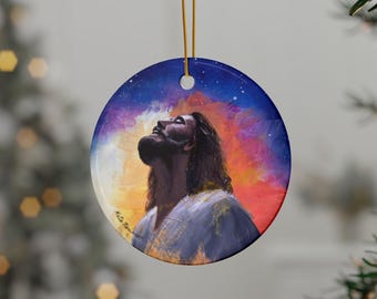 May include: Round Christmas ornament featuring a painted portrait of a man with long hair looking upwards. The background is a blend of blue, purple, orange, and red hues. A gold string is attached for hanging. The ornament is a religious themed item.