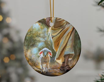 Può includere: Ornamento natalizio rotondo con un dipinto di un agnello che segue una figura in una veste. L'opera d'arte utilizza toni caldi di giallo, marrone e verde, con la firma dell'artista visibile. L'ornamento è appeso a un filo d'oro.