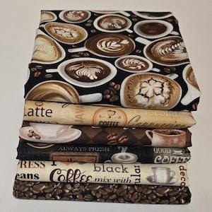 May include: A stack of fabric pieces with coffee-themed patterns. Designs include coffee cups with latte art, coffee beans, and text like "Latte," "Coffee," and "Always Fresh." The fabrics feature shades of brown, black, and beige.