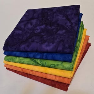 May include: A stack of colorful fabric squares in a rainbow of colors. The fabrics include shades of purple, blue, green, yellow, orange, and red. The fabric has a mottled, textured appearance.