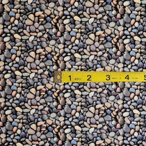 May include: A close-up of a fabric with a repeating pattern of small, smooth, gray, brown, and blue stones. A yellow measuring tape is placed on top of the fabric, showing the measurement of 1 inch.