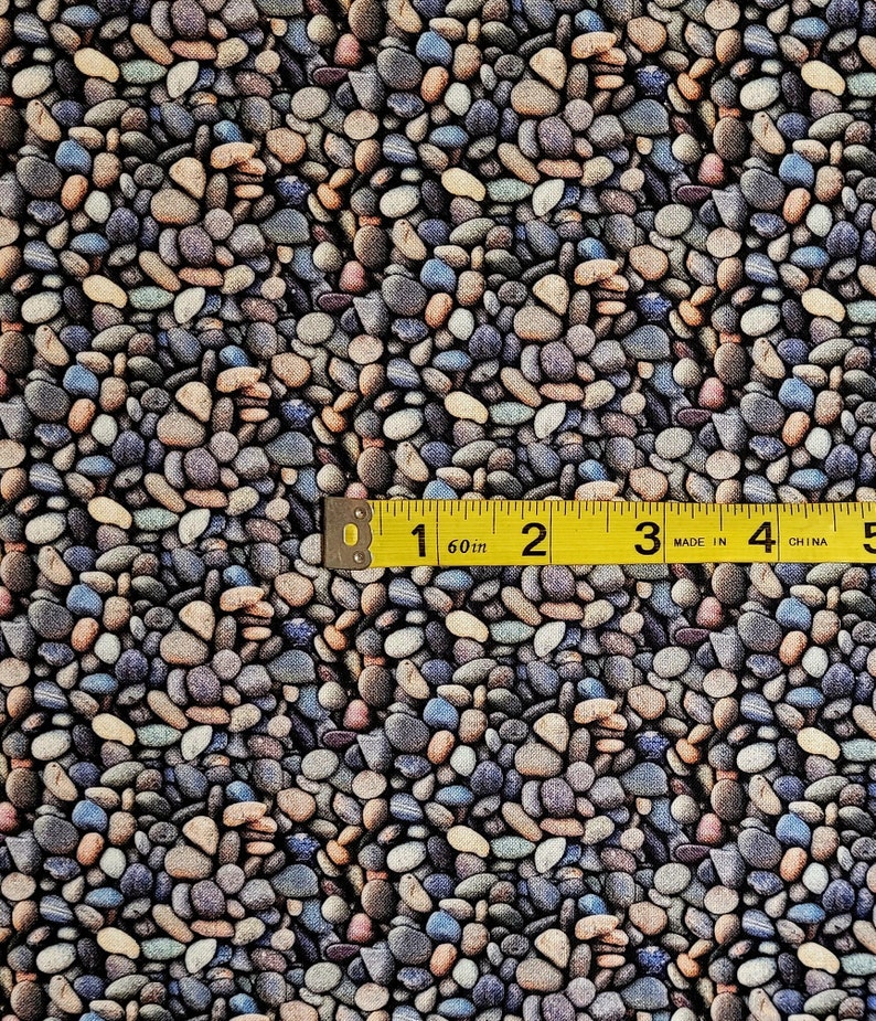 May include: A close-up of a fabric with a repeating pattern of small, smooth, gray, brown, and blue stones. A yellow measuring tape is placed on top of the fabric, showing the measurement of 1 inch.