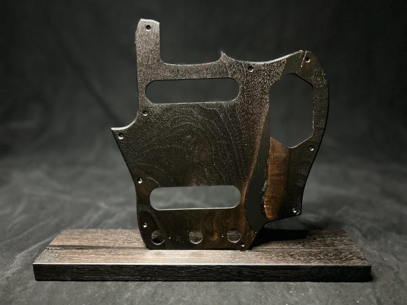 Puede incluir: Un golpeador de guitarra de madera de color marr&oacute;n oscuro con veteado, que presenta un dise&ntilde;o recortado con m&uacute;ltiples orificios para tornillos y dos ranuras rectangulares para pastillas, montado verticalmente sobre una base de madera rectangular a juego.