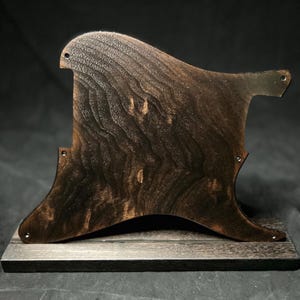 May include: A dark brown wooden guitar pickguard with a prominent, wavy grain pattern. The pickguard features several small pre-drilled screw holes along its curved edges and is mounted upright on a rectangular wooden base of the same color.