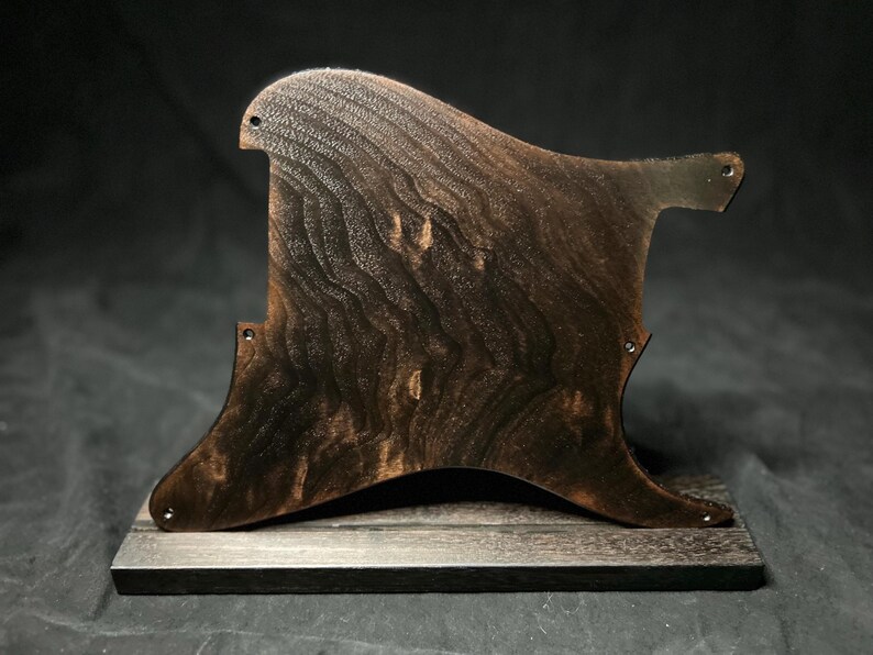 May include: A dark brown wooden guitar pickguard with a prominent, wavy grain pattern. The pickguard features several small pre-drilled screw holes along its curved edges and is mounted upright on a rectangular wooden base of the same color.