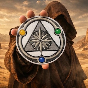 May include: A round patch featuring a compass rose design within a triangle, surrounded by a silver border. The patch has yellow, green, and blue accents. The background shows a desert landscape with mountains and a person in a brown hooded robe.