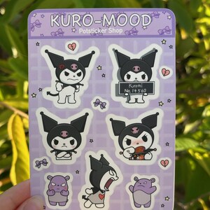 May include: A sheet of stickers featuring Kuromi, a black and white Sanrio character with a skull and crossbones on her head. The stickers are on a purple background with white stars and hearts. The text "KURO-MOOD Potsticker Shop" is at the top of the sheet. One sticker shows Kuromi holding a sign that says "Kuromi No. 14 560".