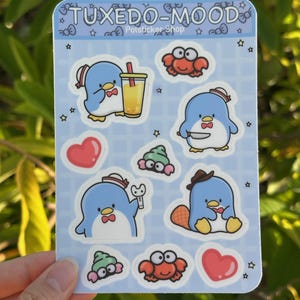 May include: A sheet of stickers featuring a blue penguin wearing a sailor hat and bow tie in various poses. The stickers also include a red crab, a green sea slug, and a heart. The sheet is on a blue background with white stars and the text "TUXEDO-MOOD" and "Potsticker Shop".